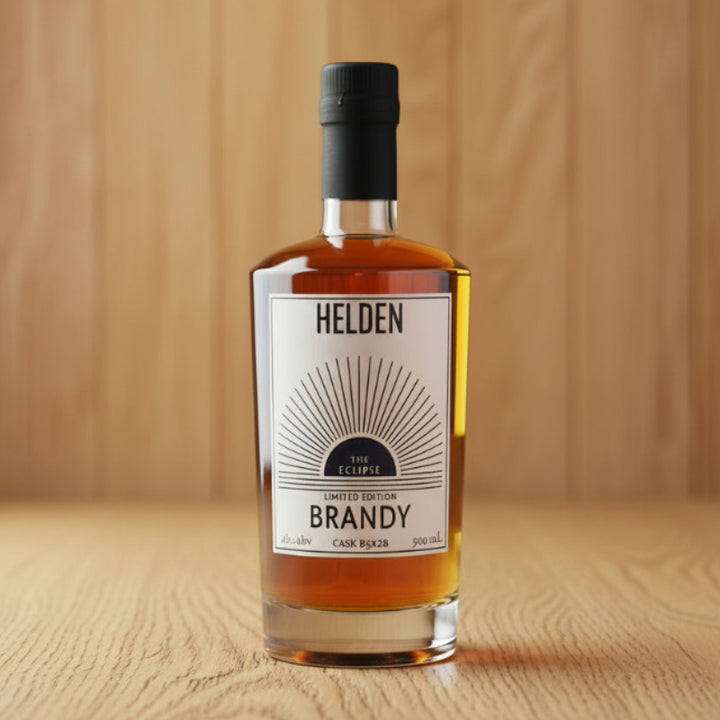 A bottle of Helden The Eclipse Limited Edition Brandy, showing the amber liquid inside a clear glass bottle with a white label and a black cap, set against a plain white background.